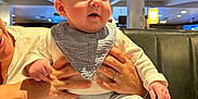 Rohan joined the competition — help win amazing prizes! baby, infant, child, person, hands, bib, clothing, socks, pants, table, wood, restaurant, indoor, lighting, face, eyes, expression, support, adult, seating