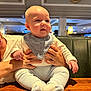 baby, infant, child, person, hands, bib, clothing, socks, pants, table, wood, restaurant, indoor, lighting, face, eyes, expression, support, adult, seating