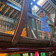Rohan is registered to the contest to win money with this photo: child, indoor_playground, netting, mesh, play_structure, blue_clothing, young_child, grip, looking_down, colorful_background, wall_painting, play_area, wooden_structure, safety_mesh, feet, shorts, toddler, playground_equipment, indoor, active