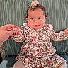 Nayla is registered to the contest to win money with this photo: baby, child, infant, portrait, sitting, smiling, floral_dress, pink_flower, hair_accessory, striped_chair, indoor, adult_hand, fashion, cute, face, eyes, dress, tights, furniture, photography