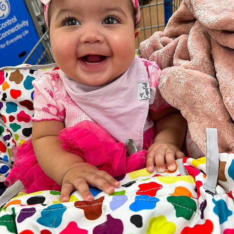 Kynsley is registered to the contest to win money with this photo: baby, blanket, bright_colors, child, clothing, colorful, cute, face, hands, happy, headband, indoor, patterned, pink_outfit, portrait, retail, shopping_cart, smiling, soft_texture, toddler