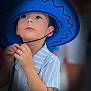 child, boy, hat, cowboy_hat, blue_hat, striped_shirt, portrait, face, curious, innocent, young_child, looking_up, hands, chin_strap, soft_background, close_up, casual_clothing, indoor, person, cute