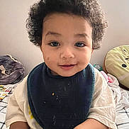 Josiah joined the competition — help win amazing prizes! baby, bed, bib, casual_clothing, child, cozy, curly, curly_hair, face, happy, home, indoor, person, pillow, shirt, sitting, skin, smile, toddler, young_child
