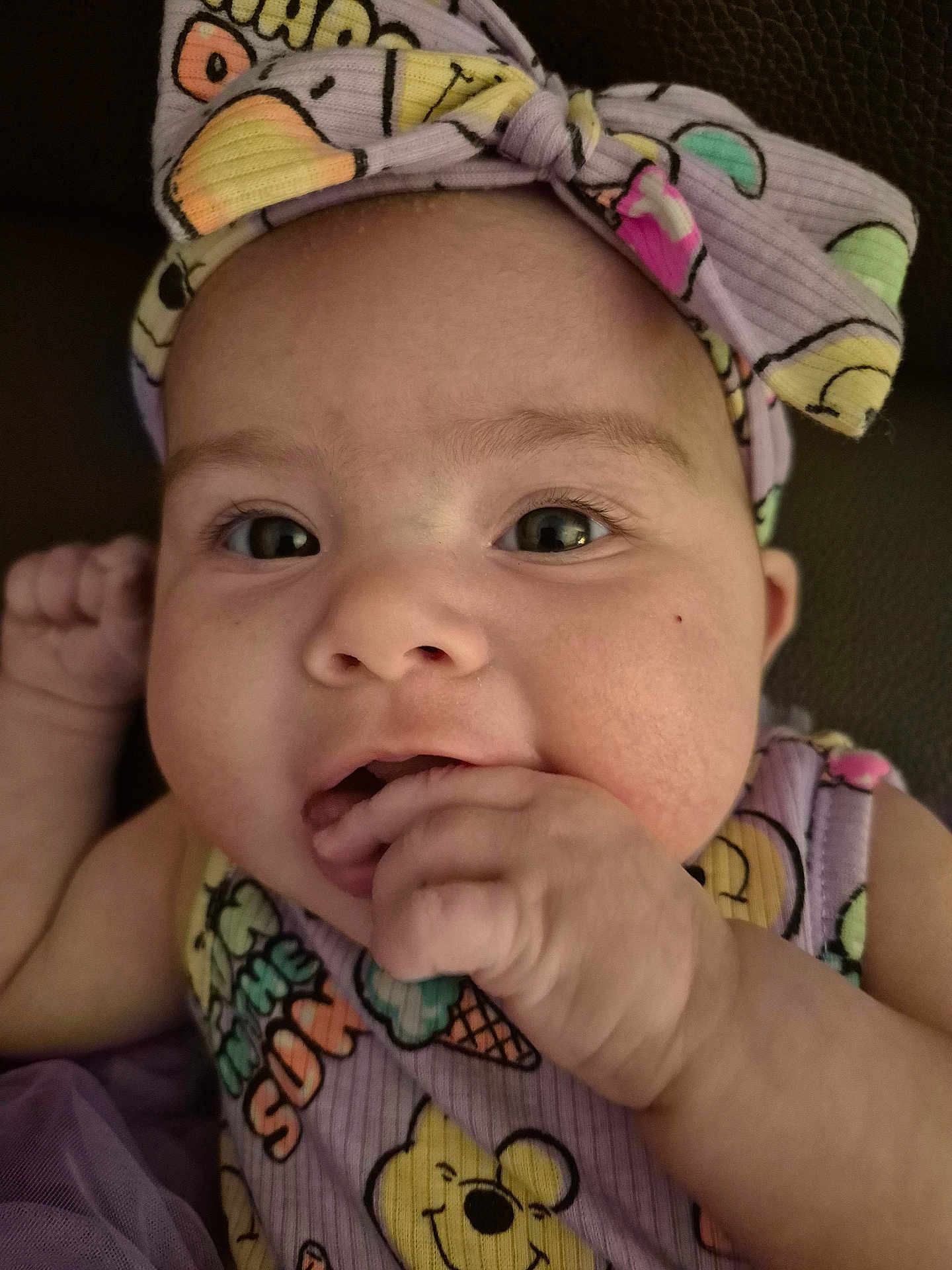 Leora joined the competition — help win amazing prizes! baby, infant, face, eyes, hand_in_mouth, headband, bow, onesie, ice_cream_pattern, cartoon_bear, close_up, portrait, cheeks, fingers, curious_expression, couch_background, purple_clothing, soft_light, newborn, adorable