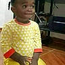 child, toddler, sitting, portrait, indoors, yellow_clothing, patterned_shirt, braids, face, side_glance, serious_expression, hands, floor, tile_floor, television, furniture, living_room, photograph, smartphone_screenshot, casual_clothing
