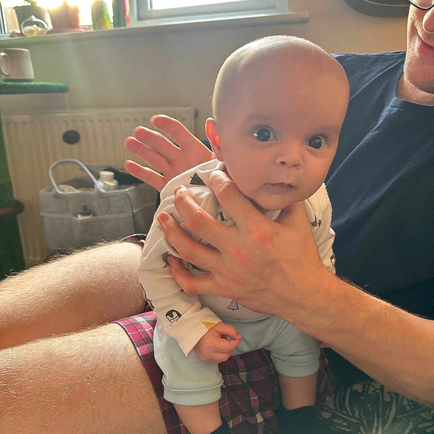 Kaii is registered to the contest to win money with this photo: adult, baby, carpet, clothing, cozy, curious, glasses, hands, home, indoor, legs, natural_light, pattern, person, plaid_shorts, portrait, radiator, sitting, socks, window