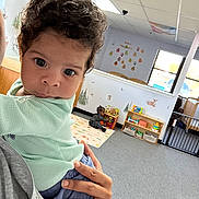 Milo joined the competition — help win amazing prizes! toddler, child, curly_hair, indoor, daycare, toys, adult_hand, carpet, window, wall_decor, sitting, person, room, play_area, curious, baby, holding, casual_clothing, floor, natural_light