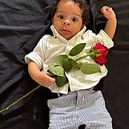 Milo is registered to the contest to win money with this photo: baby, casual, child, clothing, curly_hair, cute, dark_background, flower, green_leaf, hand, infant, laying_down, petal, plant, portrait, rose, striped_pants, white_shirt, wide_eyes, young_child