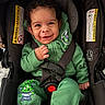 baby, car_seat, casual_wear, child, closeup, curly_hair, cute, expressive_face, green_clothing, happy, indoors, infant, playful, portrait, safety_harness, seated, smiling, warning_label, white_socks, young_child