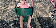 Octavia is registered to the contest to win money with this photo: toddler, child, swing, playground, outdoor, smiling, happy, green_swing, wood_chips, bench, summer, sunlight, grass, trees, person, fun, play, barefoot, casual_clothing, daytime