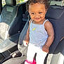 car_seat, child, curly_hair, cute, grey_tank_top, happy, indoor, jewelry, leather_seat, necklace, person, pink_hair_clips, pink_ruffles, portrait, smiling, sunlight, toddler, vehicle_interior, white_pants, white_shoes