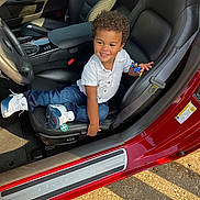 London is registered to the contest to win money with this photo: toddler, child, car, corvette, toy, smiling, curly_hair, white_shirt, jeans, sneakers, leather_seat, seatbelt, sunlight, dashboard, steering_wheel, car_interior, happy, playing, vehicle, door