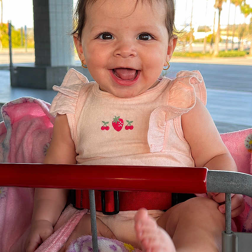 Victoria is registered to the contest to win money with this photo: baby, child, smiling, shopping_cart, blanket, pink, outdoor, daylight, happy, person, clothing, earrings, foot, seatbelt, metal, grid_floor, trees, architecture, sunlight, cute