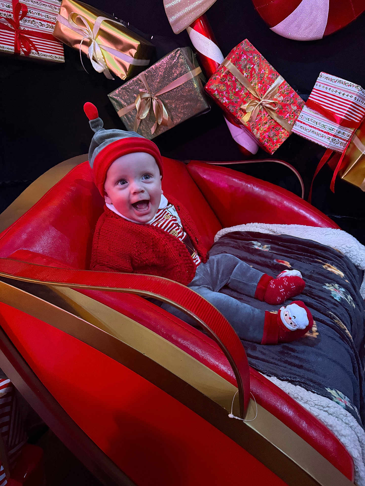 Noah joined the competition — help win amazing prizes! baby, child, smiling, red_clothing, hat, socks, sleigh, christmas, presents, gift_wrap, holiday, festive, blanket, cozy, indoor, celebration, portrait, happy, cute, seasonal