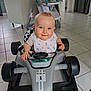 baby, bib, blue_eyes, child, cute, floor_tiles, furniture, hands, high_chair, home, indoor, infant, person, playful, race_car, seat, smiling, steering_wheel, toy_car, vehicle
