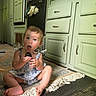 Emmie is registered to the contest to win money with this photo: baby, toddler, child, sitting, curious, kitchen, rug, floor, cabinet, wood_floor, green_cabinet, expression, open_mouth, holding_object, indoor, natural_light, clothing, person, home, fringe_rug