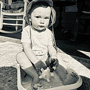 Emmie is registered to the contest to win money with this photo: toddler, child, hat, water, toy, plastic_tub, porch, rocking_chair, black_and_white, sunlight, shadow, floor, sitting, playing, outdoor, expression, clothing, barefoot, person, portrait