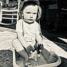 toddler, child, hat, water, toy, plastic_tub, porch, rocking_chair, black_and_white, sunlight, shadow, floor, sitting, playing, outdoor, expression, clothing, barefoot, person, portrait