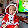 toddler, child, santa_outfit, red_clothing, hood, indoor, television, animation, bed, furniture, person, smile, happy, holiday, festive, buttons, hand, face, cozy, room