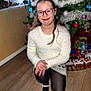 child, girl, glasses, white_sweater, black_tights, white_shoes, christmas_tree, ornaments, holiday_decorations, train_set, wooden_floor, indoor, smiling, festive, kneeled, long_hair, home, seasonal, celebration, happy