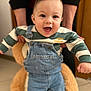 baby, child, smiling, denim_overalls, striped_sweater, plush_toy, teddy_bear, indoor, person, happy, cute, holding, floor, furniture, portrait, casual_clothing, young_child, support, hands, expression