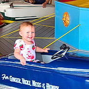 Samantha joined the competition — help win amazing prizes! toddler, child, amusement_park, car_ride, blue_car, smiling, steering_wheel, indoor, fun, playful, young_child, short_hair, casual_clothing, ride, smile, person, toy_car, family_fun, cute, happy