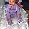baby, child, smiling, sitting, bed, bedding, pillow, blanket, purple_outfit, sneakers, curly_hair, portrait, indoors, bedroom, happy, fashion, infant, clothing, face, photography