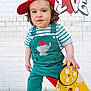 brick_wall, casual_clothing, child, cute, elephant_patch, graffiti, love_wall, outdoor, overalls, playground, playtime, red_cap, shorts, sidewalk, smiling, striped_shirt, toddler, urban, white_socks, yellow_toy