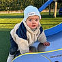 toddler, child, playground, slide, beanie, winter_clothes, jacket, outdoor, grass, fence, daylight, curious, climbing, blue_slide, green_background, shoes, person, young_child, casual_wear, park