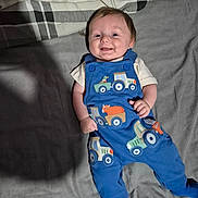 Hudson is registered to the contest to win money with this photo: baby, smiling, blue_overalls, tractor_pattern, infant, lying_down, blanket, cozy, cute, happy, child, clothing, footed_pants, indoor, portrait, young_child, toddler, casual, relaxed, soft_lighting