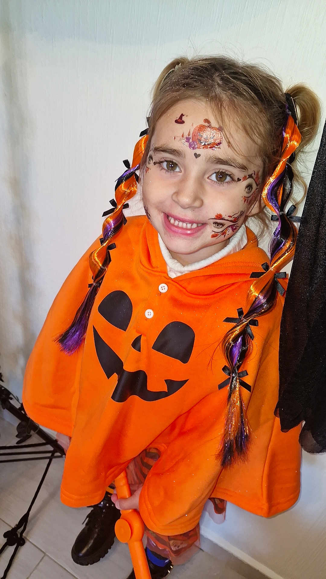 Kataleïa participe au concours pour gagner de l'argent avec cette photo : child, face_paint, halloween, costume, orange_clothing, braided_hair, smile, toy, indoor, festive, hair_ribbons, cute, happy, young, portrait, floor, black_shoes, white_wall, celebration, playful