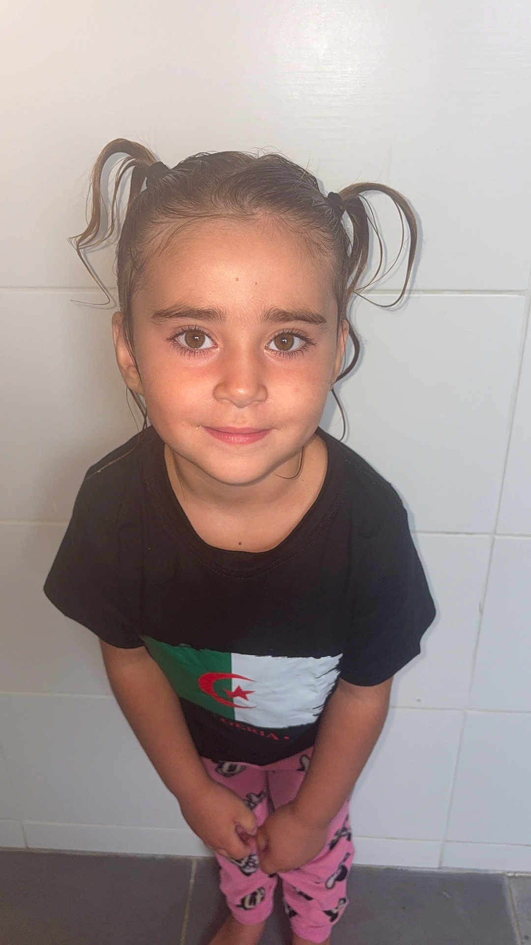 Kataleïa participe au concours pour gagner de l'argent avec cette photo : child, girl, pigtails, brown_eyes, black_tshirt, flag, pink_pajamas, standing, indoor, tile_wall, smile, cute, portrait, casual_clothing, barefoot, young, person, happy, looking_up, hair_ties