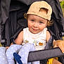 toddler, child, stroller, cap, toy, outdoor, cute, smile, curly_hair, face, baby_clothing, seatbelt, playful, sunlight, nature, hand, summer, happy, infant, park