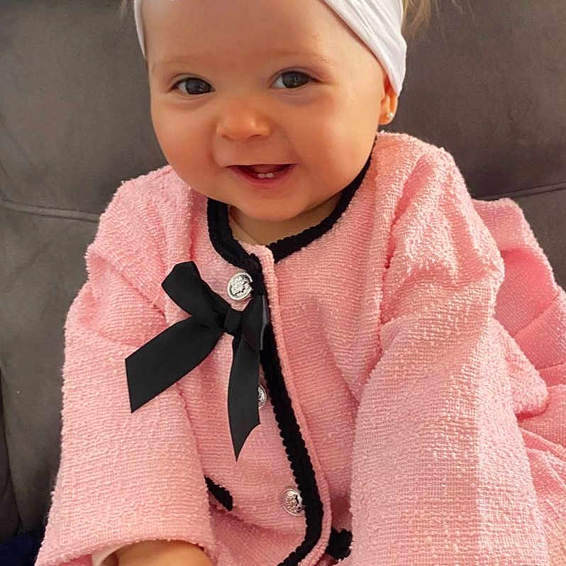 Lyana a rejoint le concours — aidez-le/la à gagner de superbes lots ! baby, black_bow, child, clothing, couch, cute, face, fashion, flower, hands, happy, headband, indoors, infant, person, pink_coat, portrait, seated, smiling, toddler