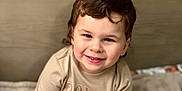 Ryett is registered to the contest to win money with this photo: child, toddler, smiling, sitting, curly_hair, blanket, wooden_blocks, age_marker, indoors, cozy, cute, happy, portrait, clothing, short_sleeve_shirt, black_pants, bedroom, soft_lighting, face, person