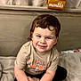 child, toddler, smiling, sitting, curly_hair, blanket, wooden_blocks, age_marker, indoors, cozy, cute, happy, portrait, clothing, short_sleeve_shirt, black_pants, bedroom, soft_lighting, face, person