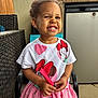 backyard, cherry_pattern, child, curly_hair, cute, daylight, earrings, furniture, happy, holding_object, minnie_mouse, outdoor, person, pink_skirt, portrait, sitting, smiling, toddler, tshirt, wicker_chair