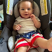 Elias is registered to the contest to win money with this photo: baby, child, car_seat, smile, shorts, american_flag, stars, stripes, hair, person, cute, indoor, clothing, seat, playful, happy, infant, leg, face, comfort