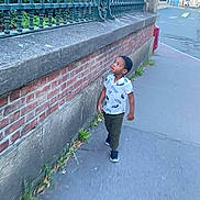 Malick Lowen a rejoint le concours — aidez-le/la à gagner de superbes lots ! child, boy, sidewalk, brick_wall, metal_fence, street, urban, outdoor, casual_clothing, short_sleeve_shirt, curious, walking, greenery, daylight, pavement, urban_environment, young_child, person, casual_shoes, summer