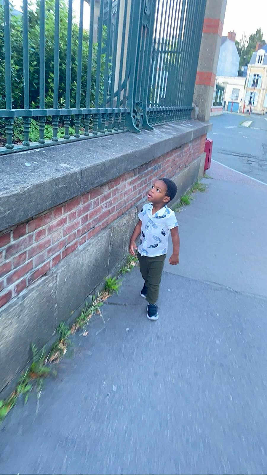 Malick Lowen a rejoint le concours — aidez-le/la à gagner de superbes lots ! child, boy, sidewalk, brick_wall, metal_fence, street, urban, outdoor, casual_clothing, short_sleeve_shirt, curious, walking, greenery, daylight, pavement, urban_environment, young_child, person, casual_shoes, summer