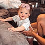 baby, child, bow, smile, couch, living_room, pillow, hand, support, happy, indoor, clothing, person, floor, footwear, home, furniture, curly_hair, skin, blue_eyes