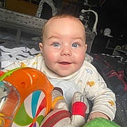 Reuben is registered to the contest to win money with this photo: baby, child, smile, blue_eyes, toy, colorful, bed, blanket, indoor, cute, happy, infant, playful, face, clothing, patterned, person, closeup, head, cheeks