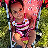 baby, child, stroller, grass, outdoor, sunlight, blue_eyes, curly_hair, pink_clothing, flower_decor, rainbow_pattern, footwear, barefoot, sitting, smiling, cute, person, summer, playful, nature