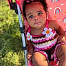 baby, child, blue_eyes, pink_stroller, rainbow_pattern, flower_applique, curly_hair, pink_clothing, outdoor, grass, drool, summer, cute, portrait, infant, seatbelt, sunlight, baby_clothes, toddler, hair_clip