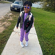 Rylynn is registered to the contest to win money with this photo: backpack, car, casual_clothing, child, daytime, fashion, grass, hair, happy, jacket, outdoor, person, purple_outfit, sidewalk, smile, sneakers, street_sign, sunglasses, tree, urban