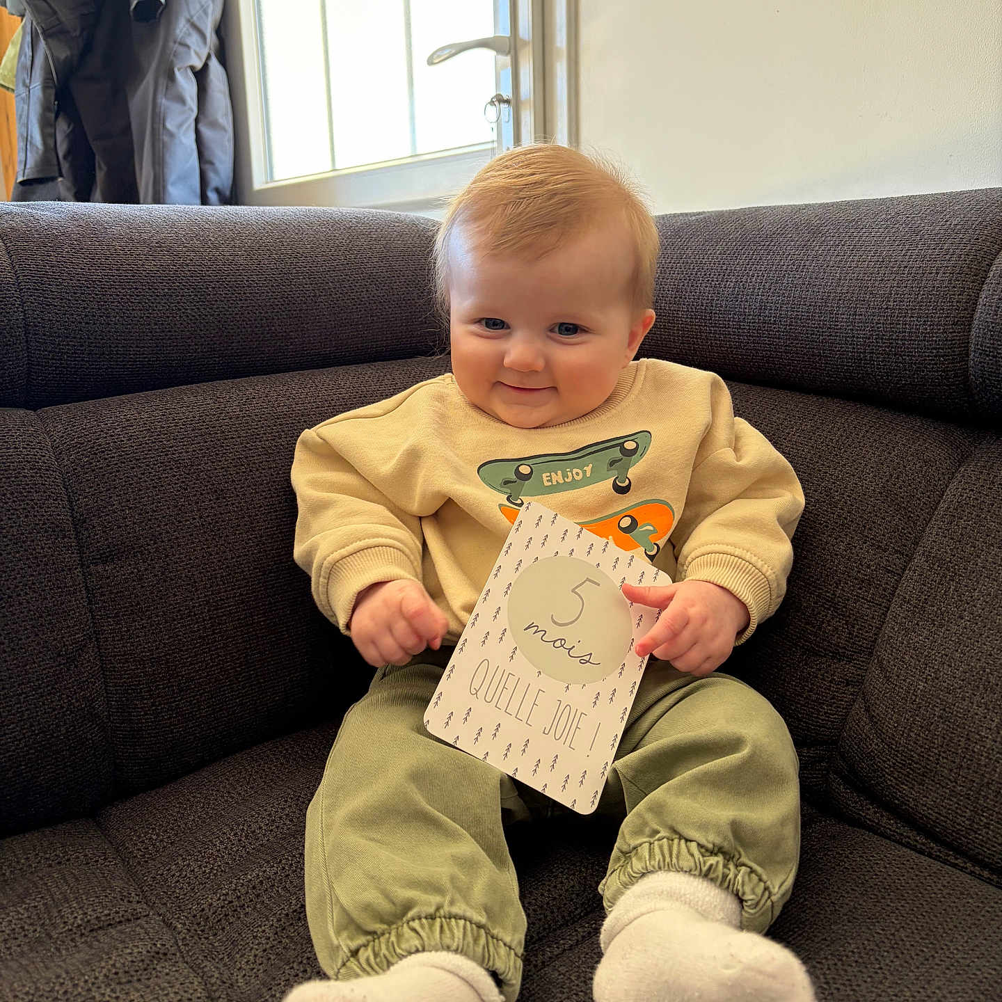 Mélio participe au concours pour gagner de l'argent avec cette photo : baby, child, coat, couch, cozy, cute, door, happy, indoor, infant, milestone_card, pants, portrait, sitting, smile, socks, sofa, soft_lighting, sweatshirt, window