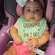 Ivy is registered to the contest to win money with this photo: baby, child, car_seat, bow, headband, bib, pink_shorts, purple_seat, cute, infant, clothing, sitting, indoors, person, expression, wide_eyes, safety_harness, seatbelt, adorable, young