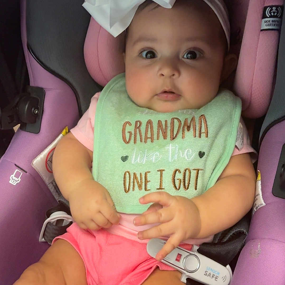 Ivy is registered to the contest to win money with this photo: baby, child, car_seat, bow, headband, bib, pink_shorts, purple_seat, cute, infant, clothing, sitting, indoors, person, expression, wide_eyes, safety_harness, seatbelt, adorable, young