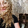 person, curly_hair, glasses, smiling, puppy, dog, brown_dog, white_chest, pet, close_up, selfie, black_shirt, wood_background, indoor, cute, young_adult, portrait, friendly, companion, animal