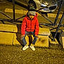 child, red_jacket, beanie, concrete_barrier, car, metal_fence, urban, night, streetlight, pavement, jeans, white_shoes, yellow_socks, sitting, outdoor, person, casual, cold_weather, expression, side_view